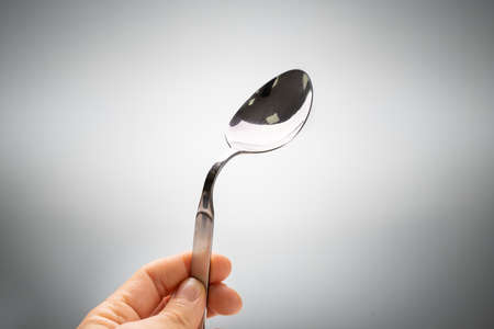 Close-up Of A Person's Hand Holding Bend Stainless Steel Spoon Against Gray Backgroundの写真素材