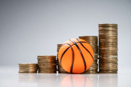 Miniature Basketball In Front Of Golden Coin Stacks On White Desk Against Gray Backgroundの写真素材