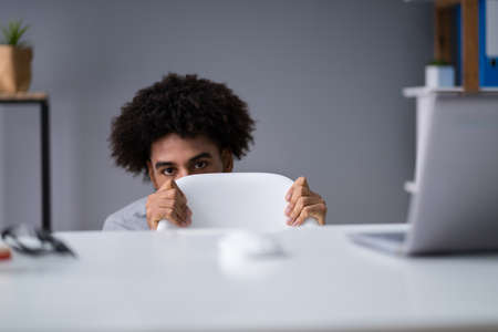 Frightened Young Businessman Hiding Behind Chair At Workplace