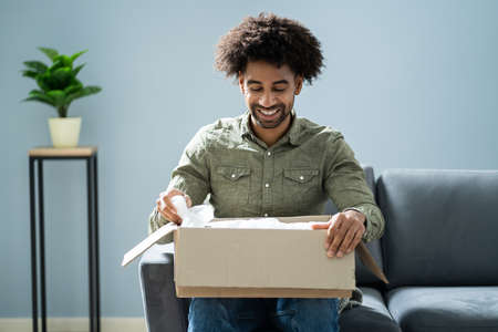 Smiling Young Man Sitting On Sofa Unpacking Received Parcelの写真素材