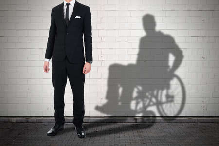 Businessman Standing Against White Brick Wall With Shadow Of A Disable Man Sitting On Wheelchairの写真素材