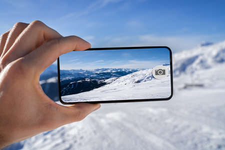 Man's Hand Taking Photograph Of Snowy Mountains On Mobile Phone