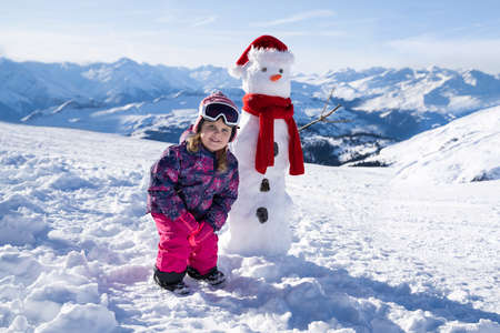 Cute Little Girl Playing Over Snowy Slope Near Snowman Looking At Cameraの写真素材