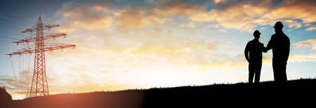 Silhouette Of Two Engineers Shaking Hands With Electricity Pylon Against Dramatic Skyの写真素材