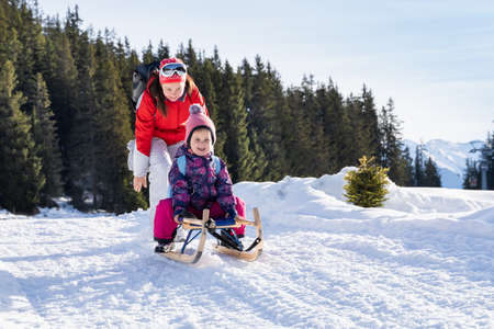 Adorable Girl Enjoying Sledding While Her Mother Pushing Sledge Against Pine Tree Forest During...