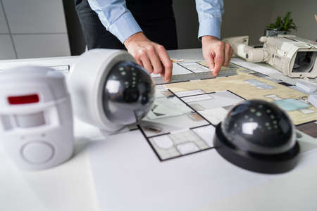 Close-up Of An Architect's Hand Drawing Blueprint With Security Equipment On Desk