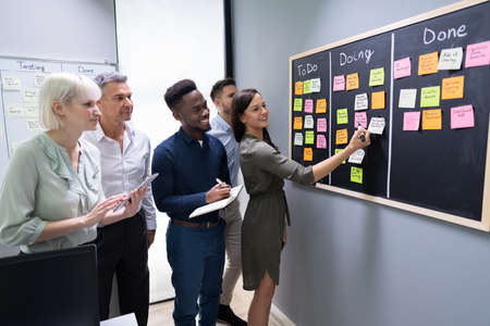 Side View Of Group Of People Writing On Sticky Notes Attached To Blackboard In Office