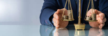 Close-up Of Lawyer's Hand Protecting Justice Scale With Coins At Table