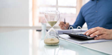 Close-up Of Hourglass In Front Of Businessperson's Hand Calculating Invoiceの写真素材