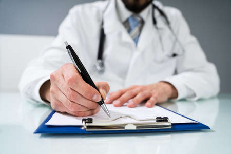Doctor's Hand Signing Document On Desk In Clinic