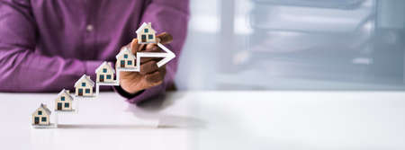 Close-up Of A Businessman's Hand Placing House Models On Increasing Blue Arrow Staircase