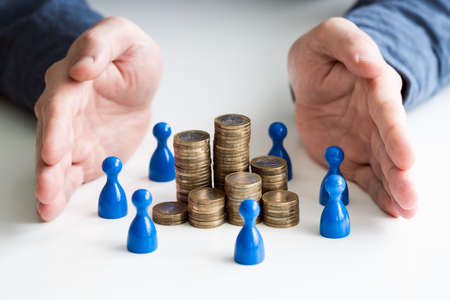 Businessperson's Hand Protecting Blue Human Figures Surrounding Stacked Golden Coins Over Deskの写真素材
