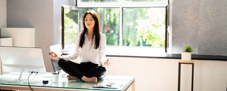 Young Businesswoman Sitting On Desk Doing Yoga In Officeの写真素材