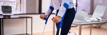 Worker With Crutches At Workplace Or Office. Handicap Rehabilitation Benefitsの写真素材