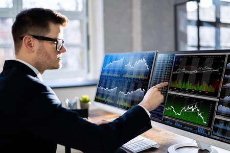 Young Male Stock Market Broker Looking At Graph On Computer At Workplace