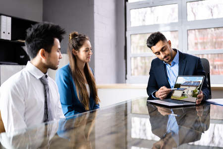 Male Architect Showing House Model On Laptop To Couple In Officeの写真素材