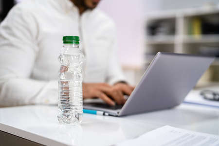 Water Bottle On Desk And Man In Foreground Using Computerの写真素材