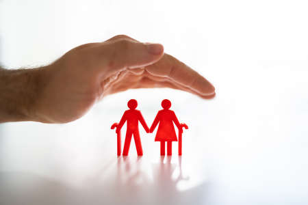 Hand Protecting Senior Couple Cutout Figures Over White Desk