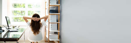 Rear View Of Relaxed Businesswoman With Hands Behind Head Sitting In Officeの写真素材