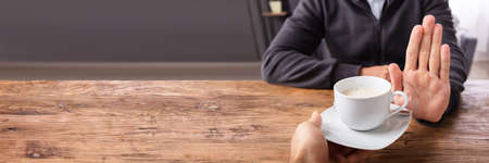 Close-up Of A Man's Hand Refusing Cup Of Coffee Offered By Person Over Wooden Deskの写真素材