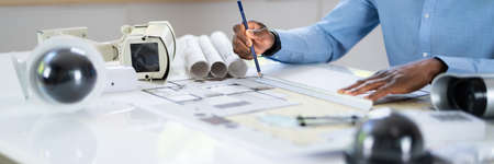 Close-up Of An Architect's Hand Drawing Blueprint With Security Equipment On Desk