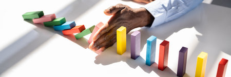 Close-up Of A Businessman Hand Stopping Colorful Dominoes From Falling On Office Deskの写真素材