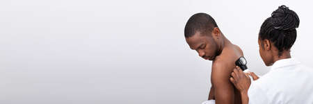 Young African Female Doctor Examining Pigmented Skin On Man's Back With Dermatoscope
