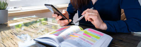 Close-up Of A Businesswoman Holding Cellphone Writing Schedule In The Diary With Pen