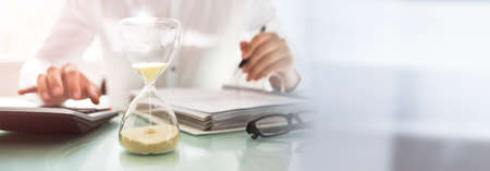 Businessman calculating bill behind sand running through the bulbs of an hourglass