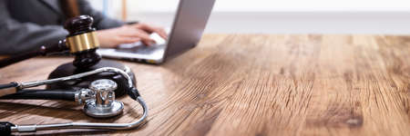 Close-up Of Mallet And Stethoscope On Gavel Over The Office Desk With Businessperson Using Laptop