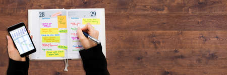 High Angle View Of A Businessperson With Mobile Phone Writing Schedule In Diary