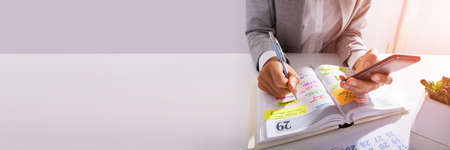 Midsection Of Person Hands With Mobile Phone And List Of Work In Diary On Desk