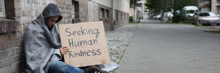 Male Beggar In Hood Showing Seeking Human Kindness Sign On Cardboardの写真素材