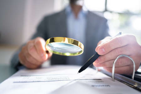 Close-up Of A Businessperson's Hand Looking At Receipts Through Magnifying Glass At Workplaceの写真素材