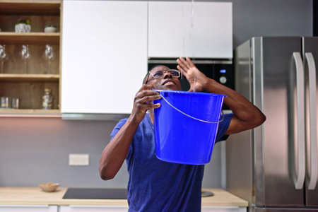 Worried Man Holding Bucket While Water Droplets Leak From Ceiling In Kitchenの写真素材