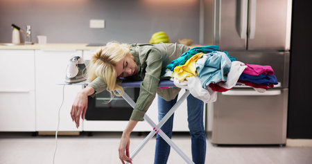 Tired Woman Sleeping On Ironing Board Next To Pile Of Clothes At Homeの写真素材