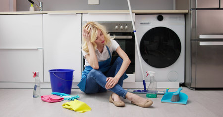 Frustrated Young Woman Sitting On Kitchen Floor And Looking At Cleaning Products At Home