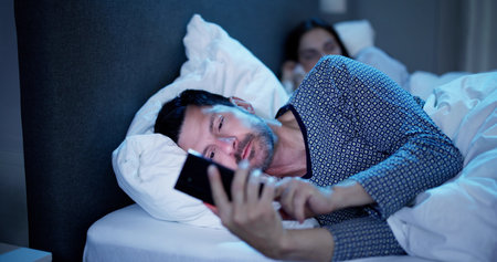 Young Man Using Cellphone While Her Wife Sitting On Bed At Nightの写真素材