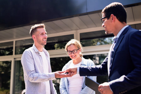 Young salesman selling a new car to a couple by handing over the key at a dealership. Business in...