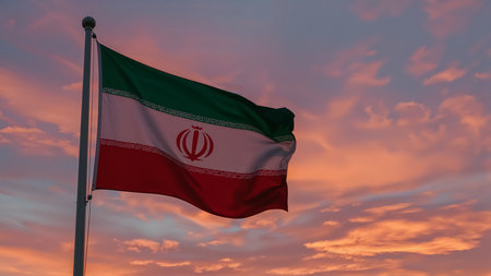 Image is a photograph featuring the national flag of Iran prominently displayed against a dramatic sunset sky. The flag is rectangular, with three horizontal bands of green, white, and red. In the center of the white band is the emblem of Iran, a stylized red tulip. The flag is attached to a white flagpole on the left side of the image. The sky in the background is filled with vibrant hues of orange, pink, and purple, creating a striking contrast with the colors of the flag. The overall composition highlights the flag's movement as it waves in the wind, with the sunset providing a vivid and colorful backdrop.の素材