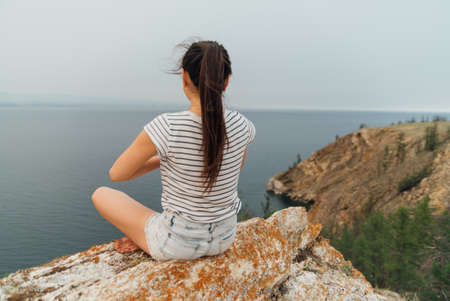 Beautiful Asian woman meditating outdoors on the rock at meadow against blue sky. the girl sits and meditates on a rock near lake Baikal. Summer yoga practice in nature. Alone.の写真素材