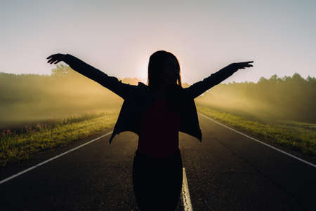 Happy woman standing with hands up on long straight road facing the sun. Sunset skyの写真素材
