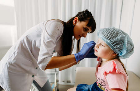Charming little girl having an ear piercing process with special equipment in a beauty center by a...