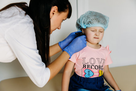 Charming little girl having an ear piercing process with special equipment in a beauty center by a professional medical workerの写真素材