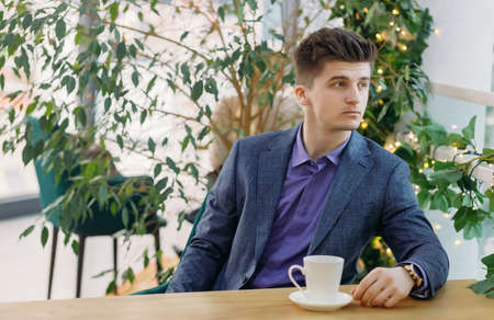 Portrait of handsome businessman in blue suit looking to the side and holding mug with fresh morning coffee while sitting at table in coffee or restaurant ...の写真素材