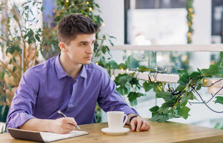 Young pensive man in a shirt with his sleeves rolled up, thinks about the project and makes notes in a notebook, drinks coffee while sitting in the office or coffee and looks awayの写真素材