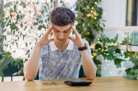 Worried male employee holding his hand behind his head and looking at the desk where the calculator and money lie while sitting at the table in the office room ..の写真素材