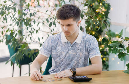 Serious young man checks bills, uses a calculator, sits at a wooden desk, accountant calculates company expenses for a financial report, plans a budget.の写真素材