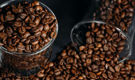 Glass transparent cup full of brown coffee beans, many coffee beans on a black background. Close-up....