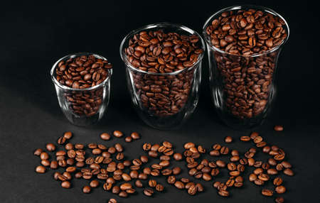 Three glass cups, overflowing with roasted coffee beans, stand next to each other. Scattered coffee...